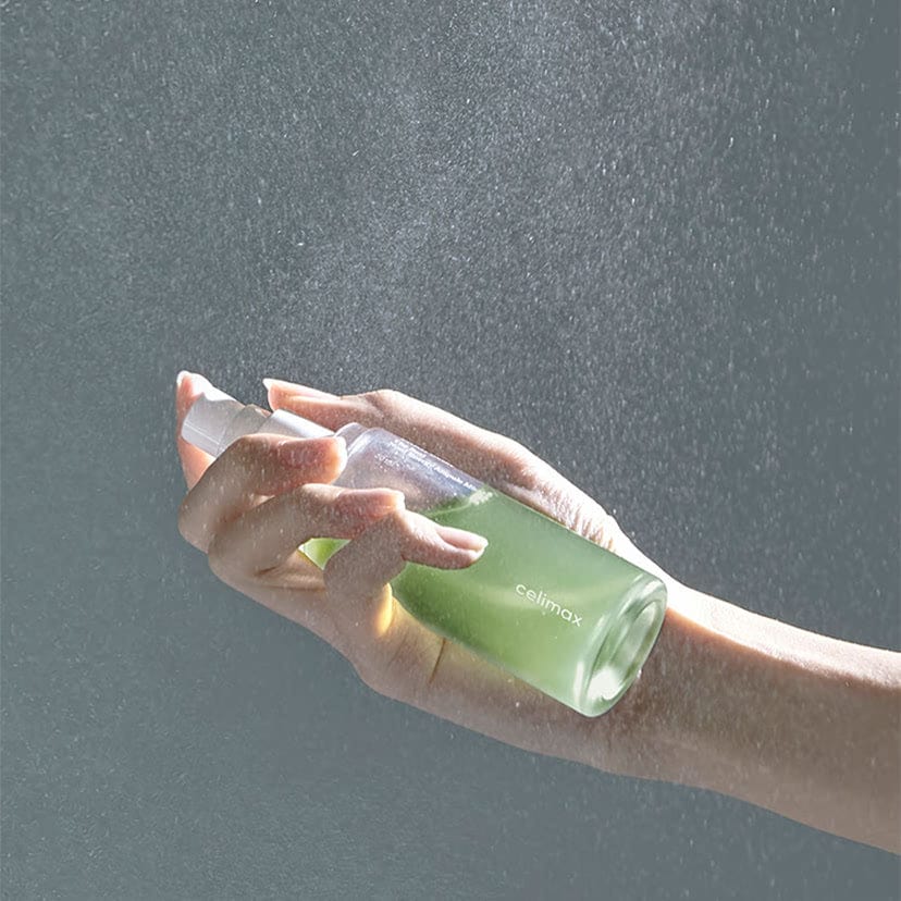 Mano pulverizando CELIMAX The Real Noni Energy Ampoule Mist de 50 ml desde su frasco verde.