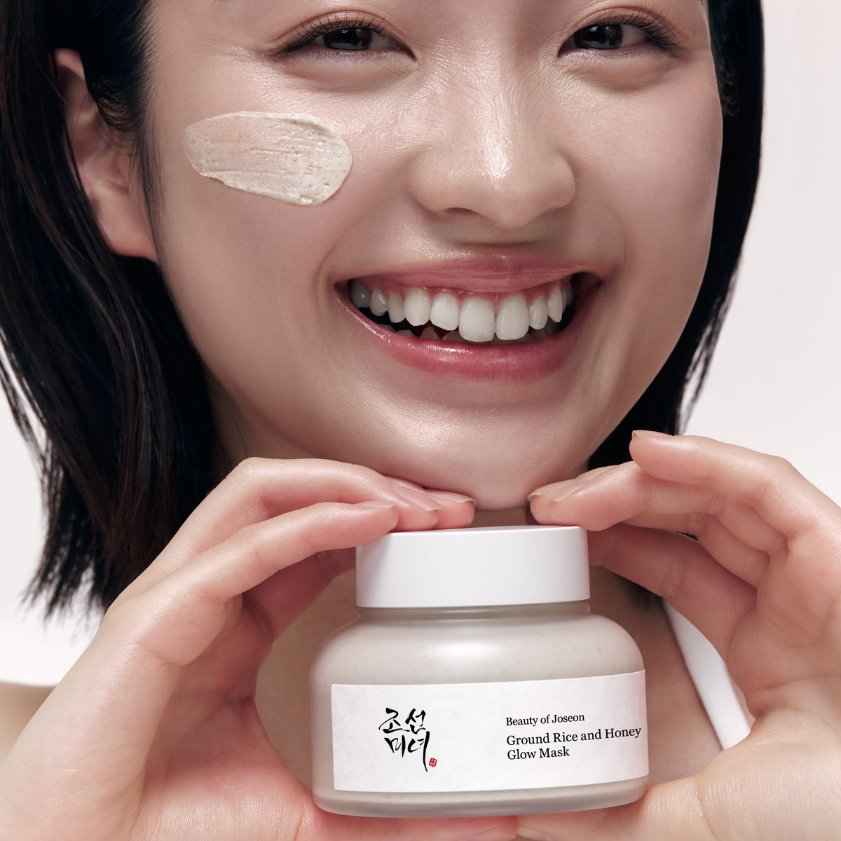 Modelo sosteniendo Beauty of Joseon Ground Rice and Honey Glow Mask de 150 ml con una pequeña cantidad de mascarilla en la mejilla