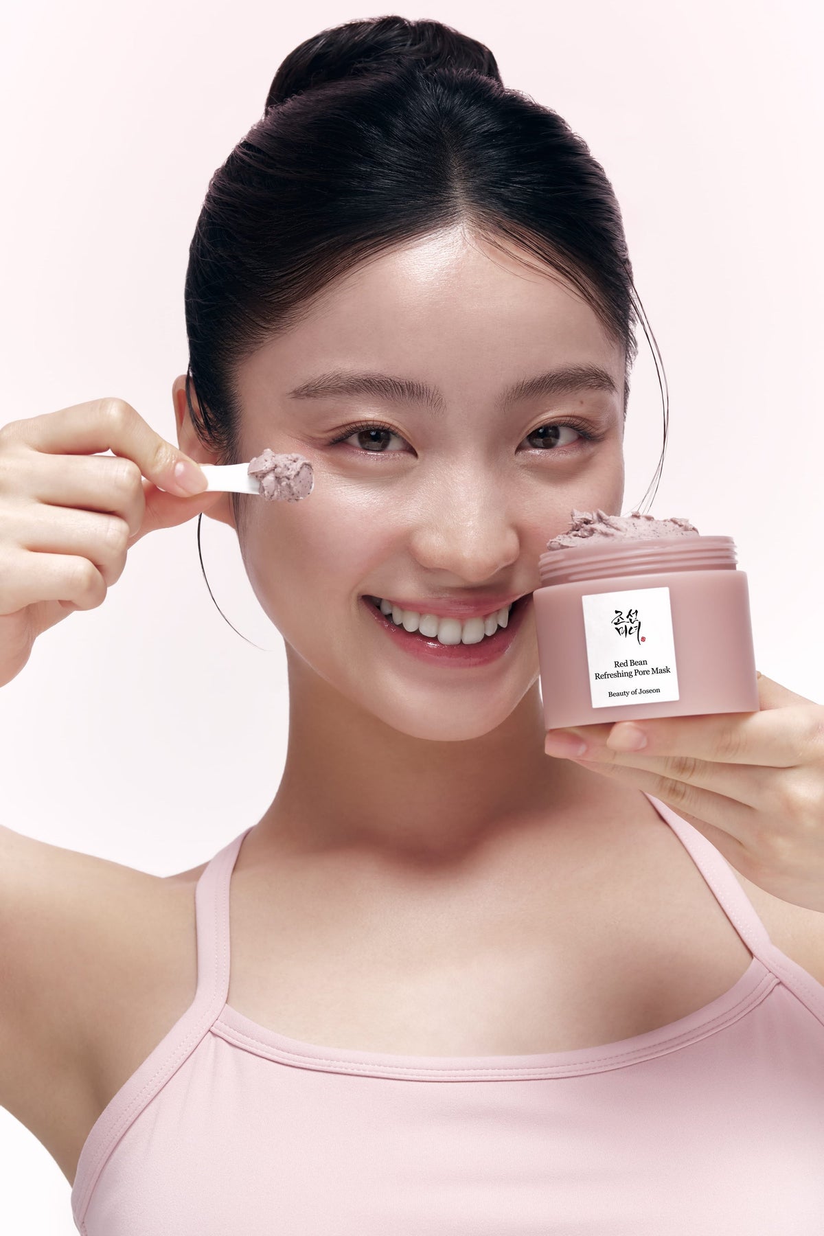 Modelo sosteniendo el tarro abierto de Beauty of Joseon Red Bean Refreshing Pore Mask de 140 ml y una pequeña espátula junto a la mejilla